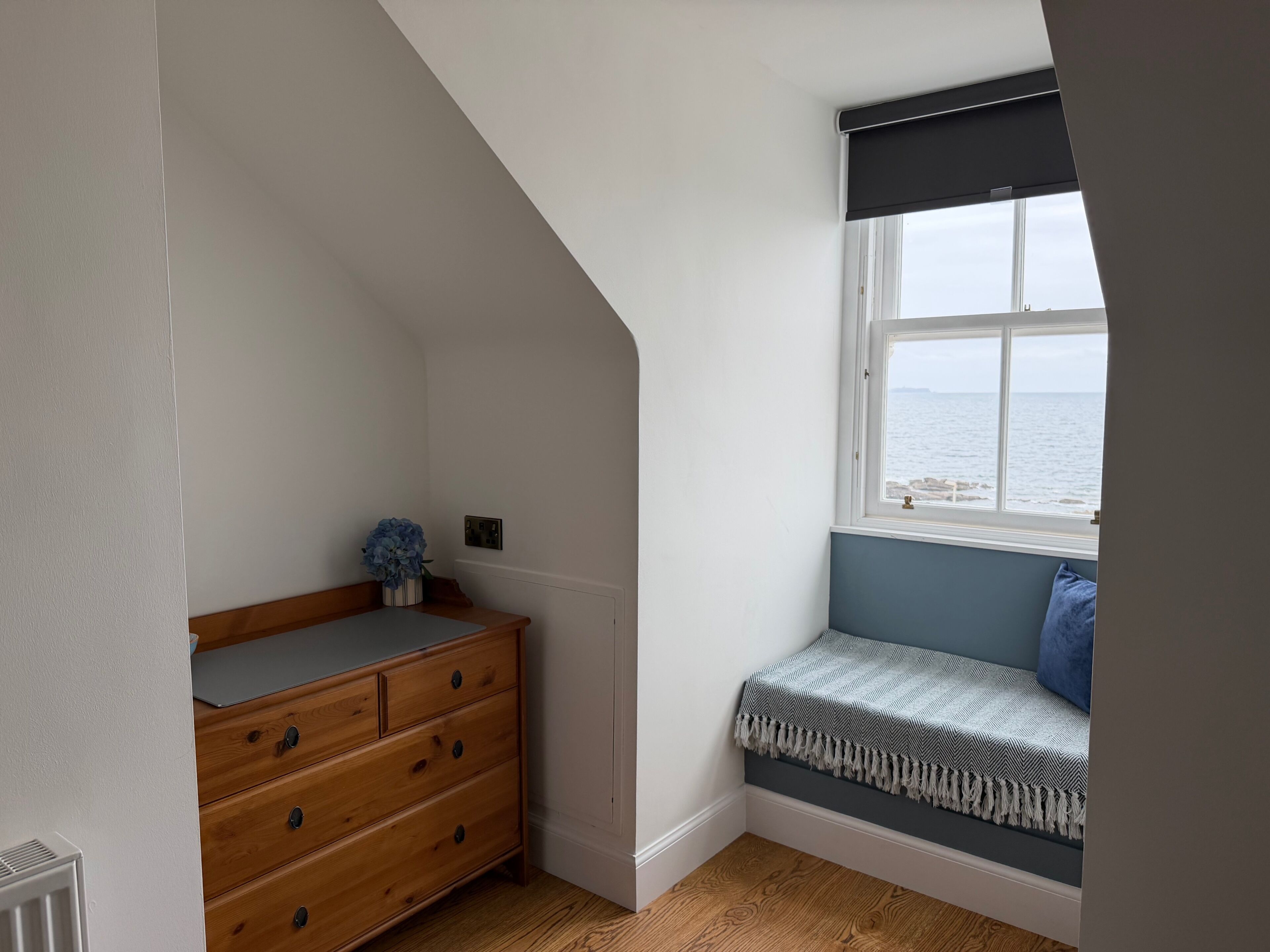 Quarto com cômoda de madeira, um banquinho com manta estampada e janela com vista para o mar.