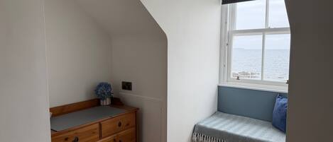 A room with a wooden dresser, a bench with a patterned throw, and a window overlooking the sea.