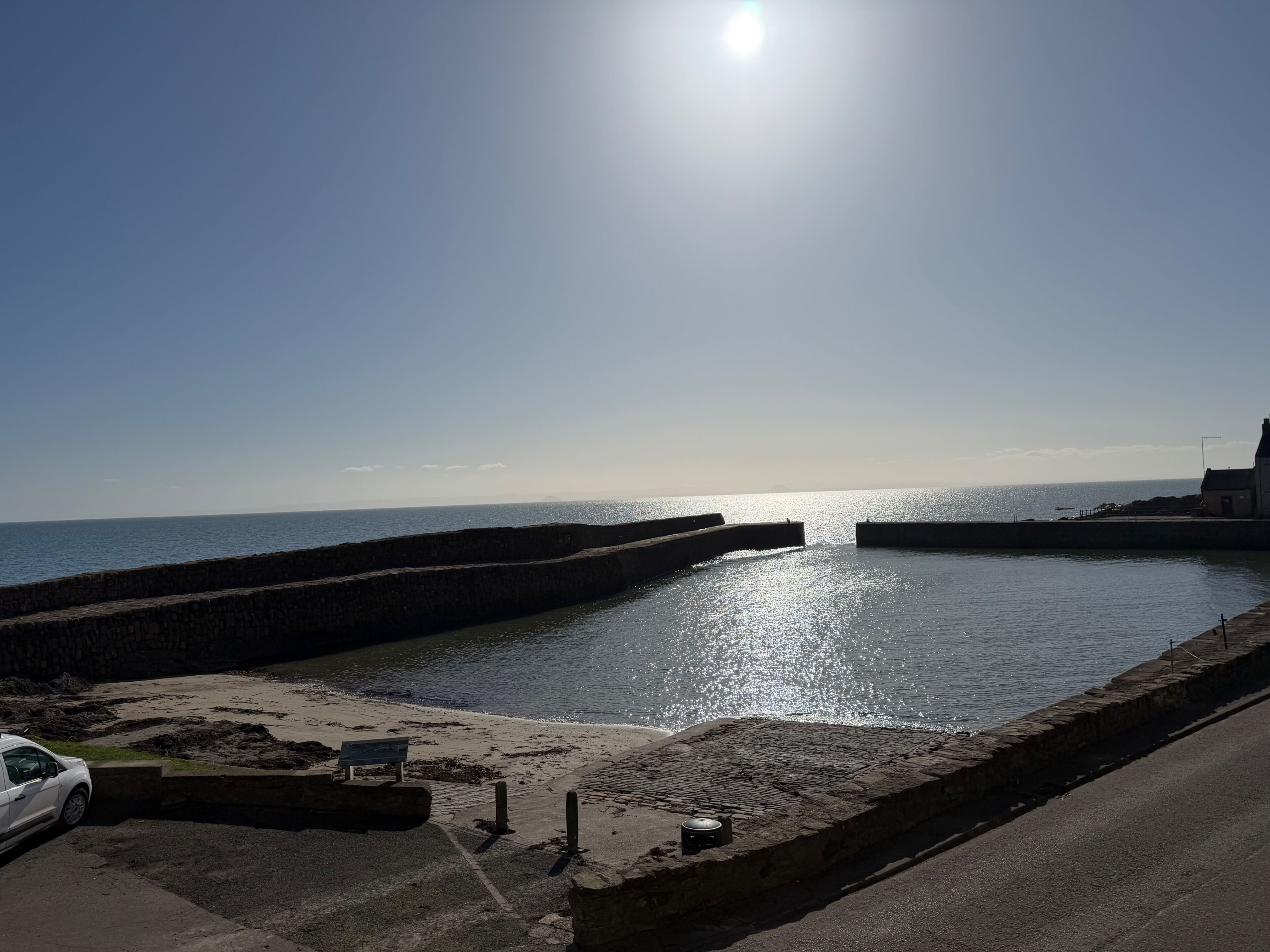 A coastal scene with a clear sky, a calm sea, and a paved walkway leading to a sandy area with a bench and a car.