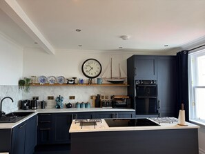 Une cuisine moderne avec des meubles de couleur sombre, un îlot central, une horloge et une fenêtre avec des rideaux.