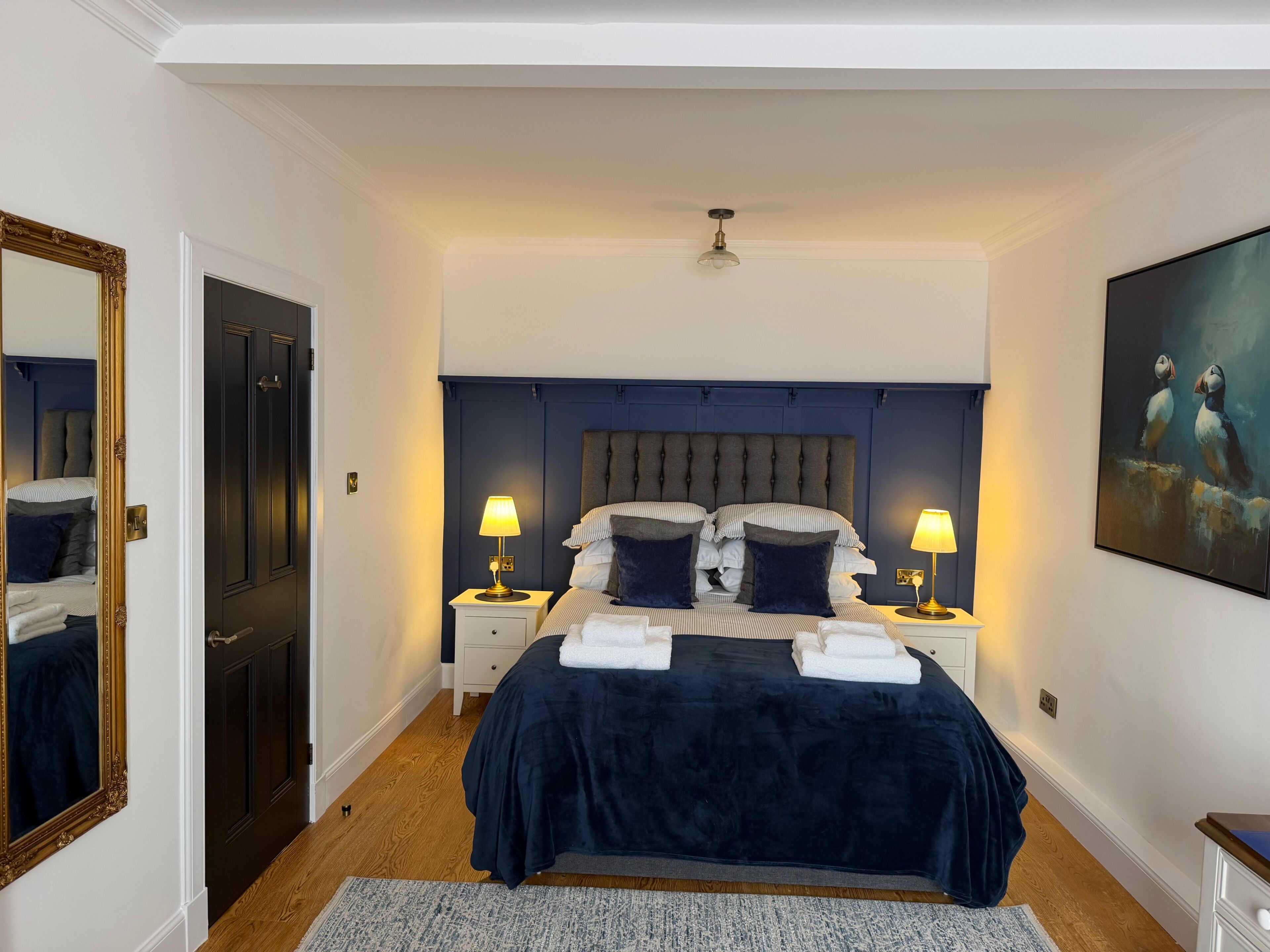 A bedroom with a dark blue headboard, two bedside tables with lamps, a large mirror, and a painting on the wall.