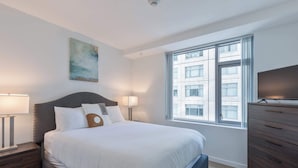 A modern bedroom with a large bed, bedside tables, a dresser, a TV, and a window with a city view. - Luxury Apt Near Kendall Square (Cambridge, MA)