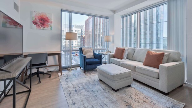 A modern living room with a sofa, ottoman, and a desk with a chair. There are large windows providing a view of the city. - Luxury Apt Near Kendall Square (Cambridge, MA)