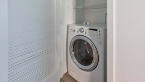 A compact, white laundry room with a front-loading washing machine, a small shelf above it, and a door with horizontal blinds. - Luxury Apt Near Kendall Square (Cambridge, MA)