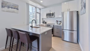 A modern kitchen with white cabinetry, stainless steel appliances, and a large island with bar stools. - Luxury Apt Near Kendall Square (Cambridge, MA)