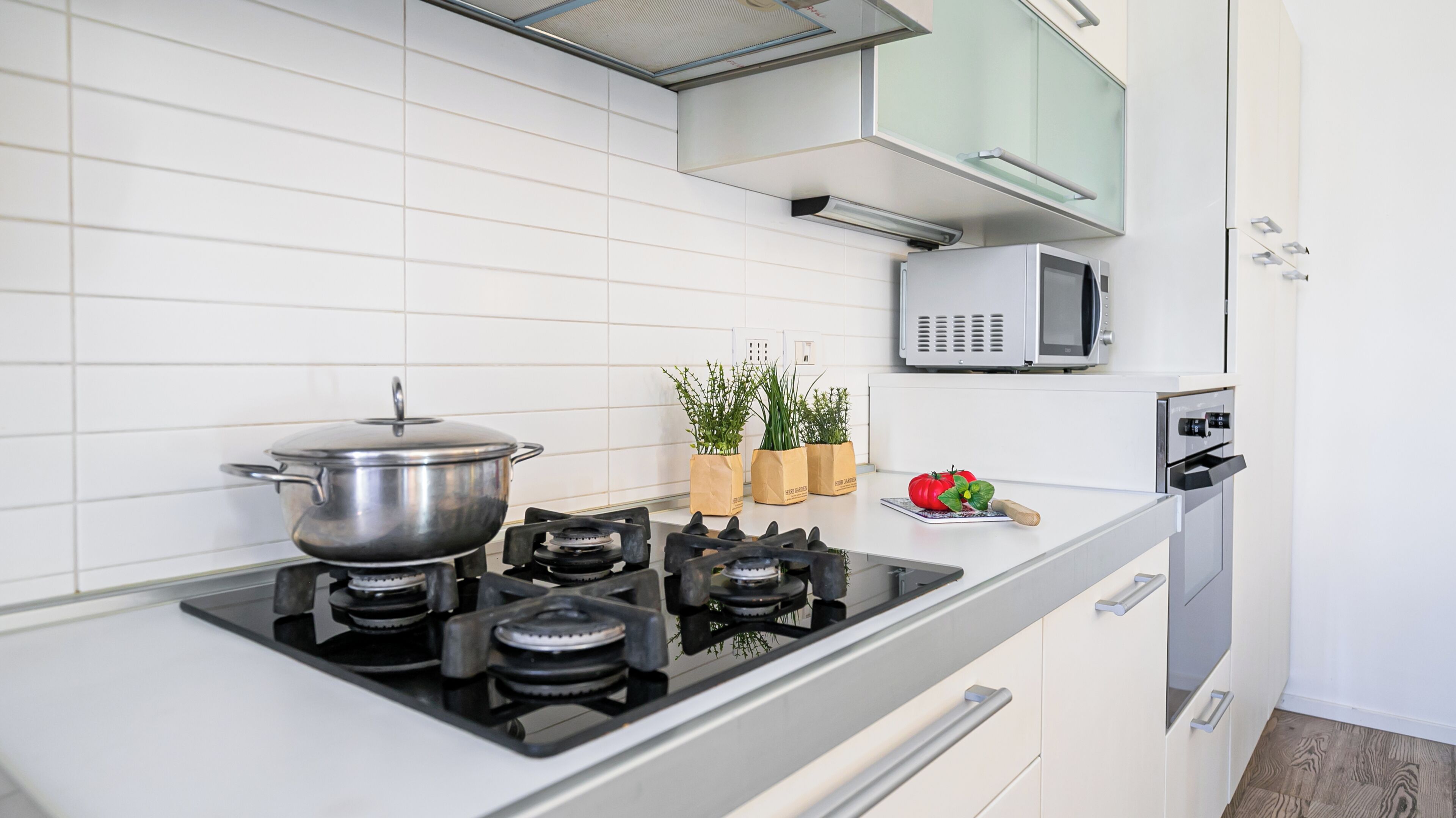Uma cozinha moderna com fogão a gás, micro-ondas e forno embutido. Há plantas em vasos sobre o balcão e um tomate vermelho sobre uma tábua de corte.
