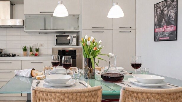 Een moderne keuken met witte kastjes, een magnetron, een fornuis en een eethoek met een glazen tafel gedekt voor twee personen. Er zijn twee hangende pendellampen, een vaas met bloemen en een poster aan de muur.