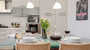 A modern kitchen with white cabinets, a microwave, a stove, and a dining area with a glass table set for two. There are two hanging pendant lights, a vase with flowers, and a poster on the wall. - Italianway Comfort - Decembrio 19 (Milan)