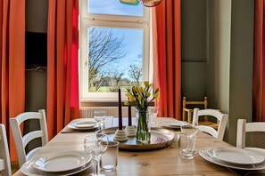 A dining table set with plates, glasses, and a vase with flowers, with a window and curtains in the background.