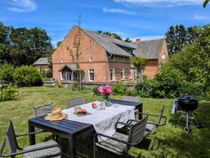 A patio with a table set for dining, surrounded by chairs, a grill, and a brick house in the background. - Himmelblau Loft with Garden – 1870 Uckermark (Nordwestuckermark)