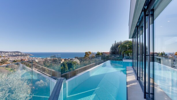 A modern infinity pool with clear blue water, surrounded by glass railings, offering a view of a coastal cityscape. - Villa Alba VI4498 by Riviera Holiday Homes (Niza)