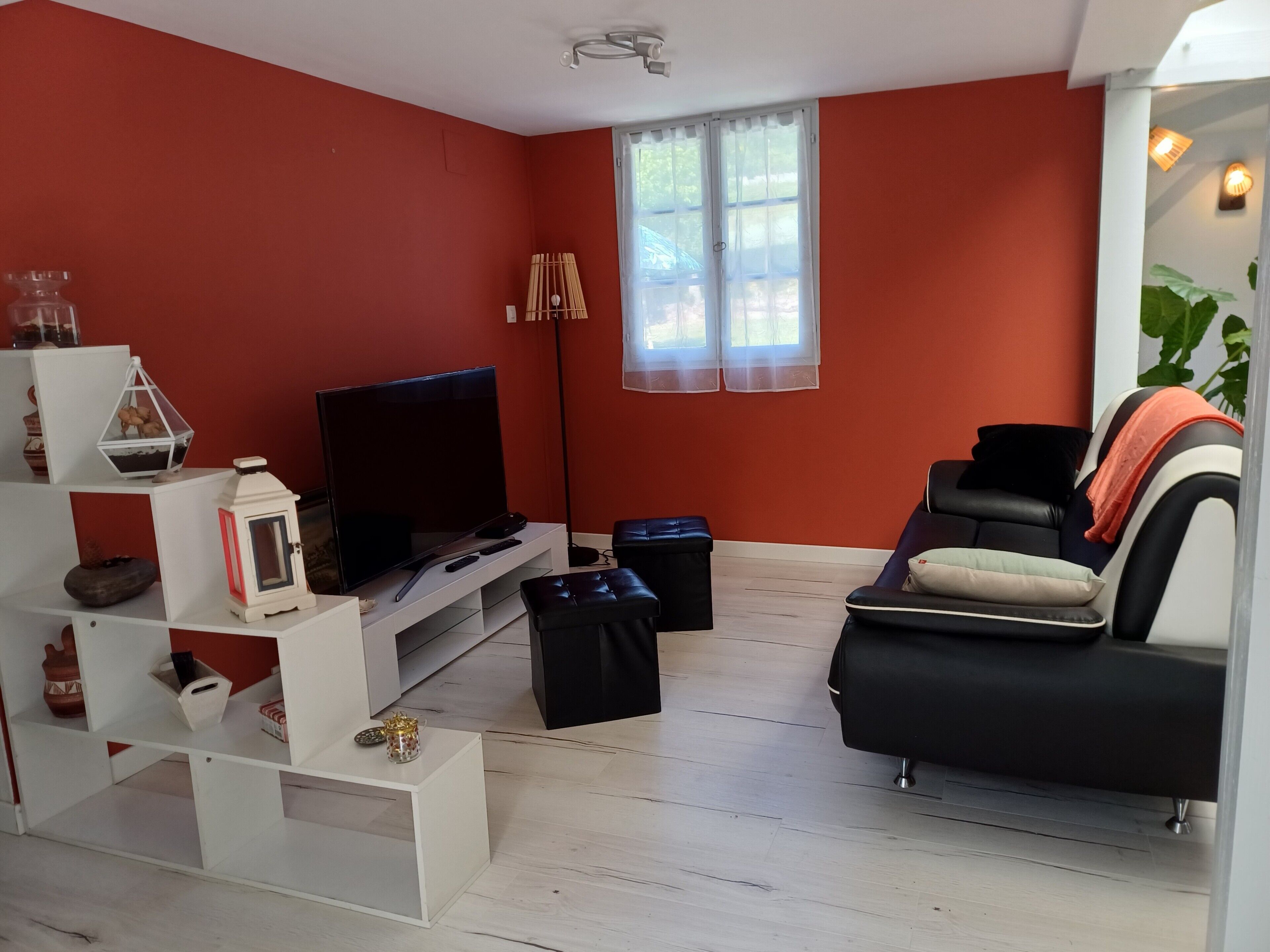 Een woonkamer met een flatscreen televisie, een zwarte leren bank, een witte tv-meubel en een raam met dunne gordijnen.