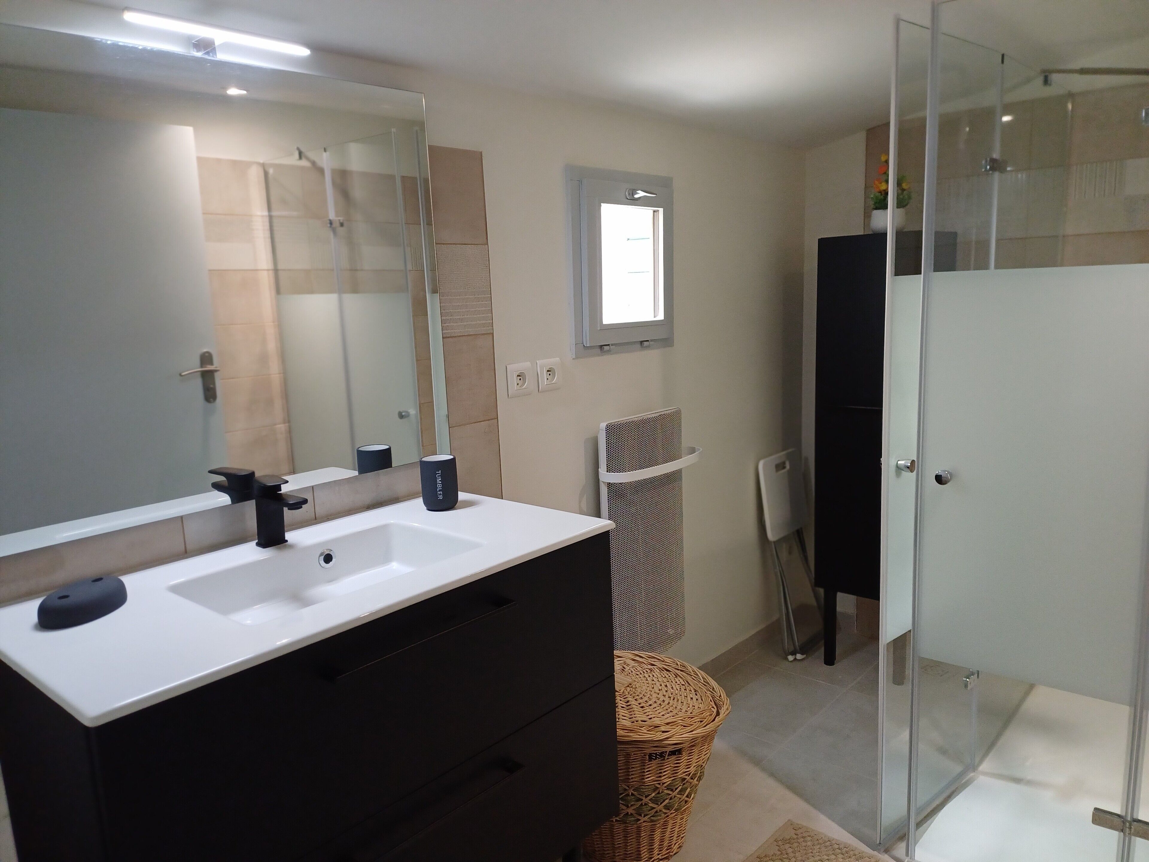 Une salle de bain avec un lavabo blanc, un grand miroir, un espace douche et une petite fenêtre.