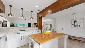 Une cuisine moderne avec des meubles blancs, un îlot central en bois, des luminaires suspendus et une vue sur l’espace de vie.