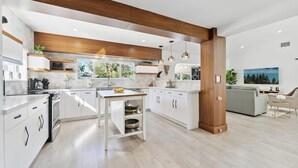 Une cuisine moderne avec des meubles blancs, un îlot central, des luminaires suspendus et une vue sur l’extérieur.