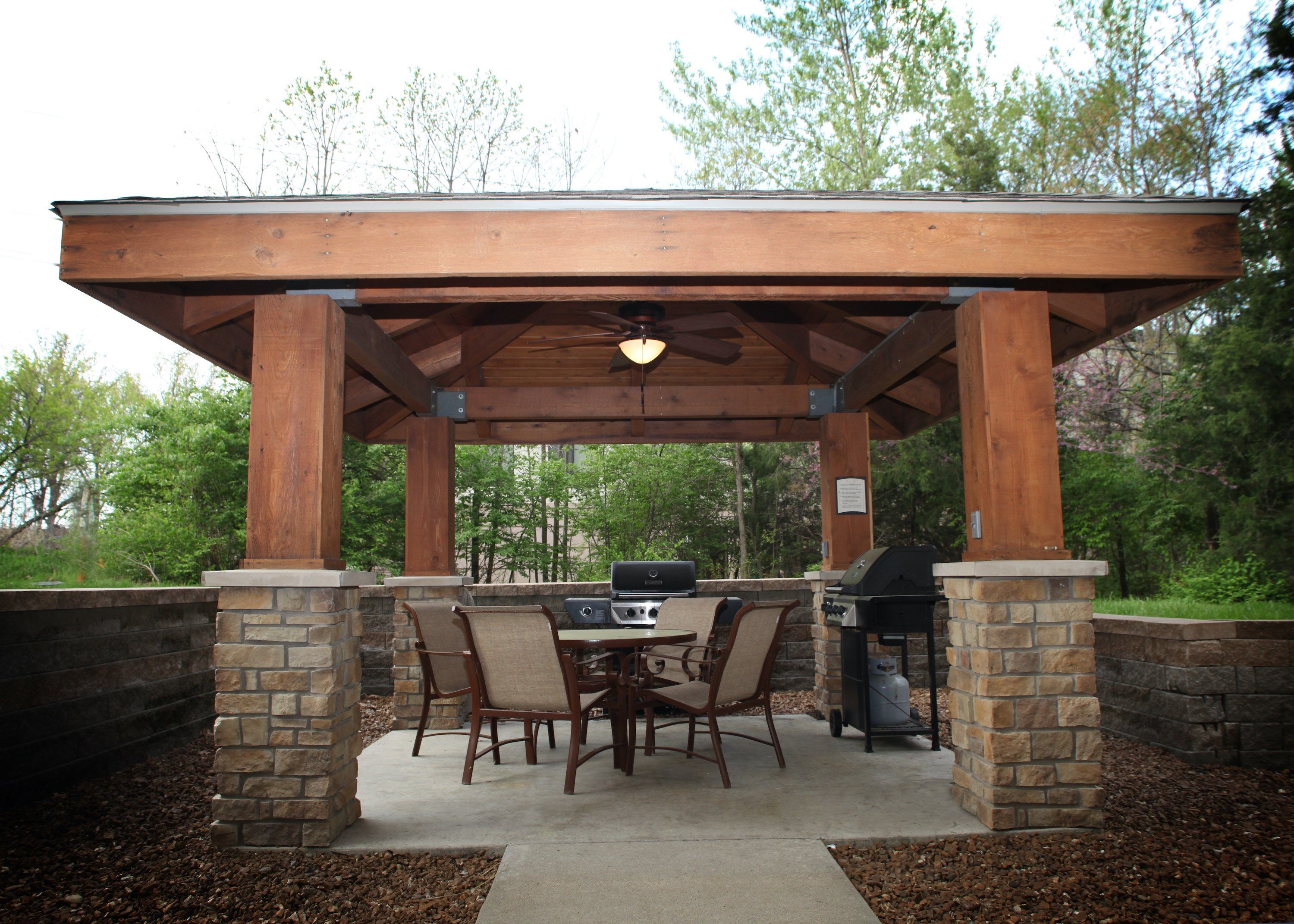 Une terrasse extérieure dotée d’une pergola en bois, de piliers en pierre et d’un espace barbecue.