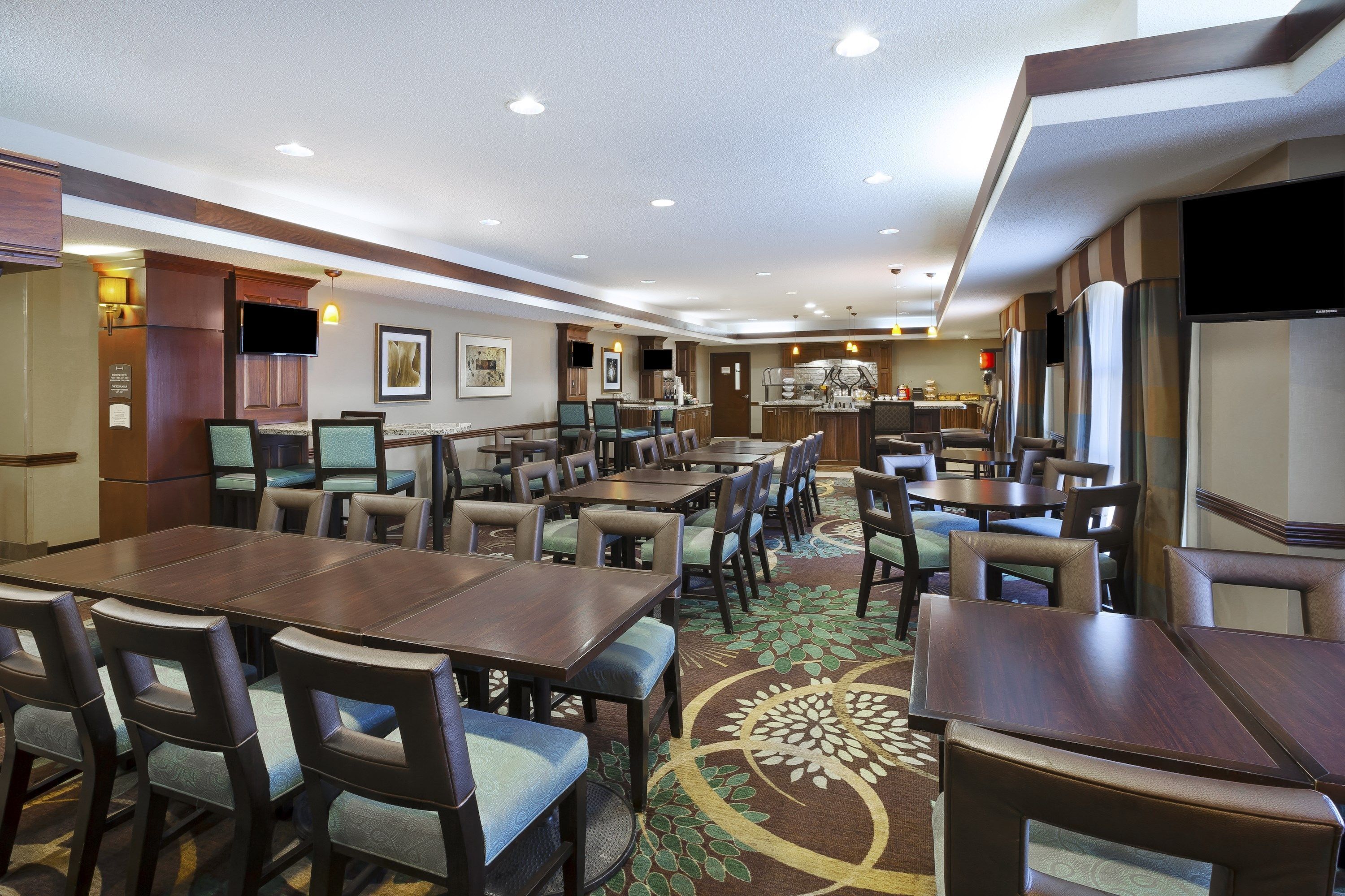 A hotel dining area with multiple tables and chairs, a kitchen in the background, and a flat-screen TV mounted on the wall.
