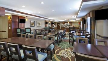 A hotel dining area with multiple tables and chairs, a kitchen in the background, and a flat-screen TV mounted on the wall.