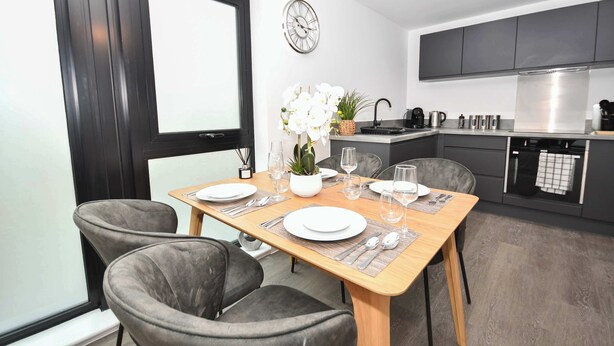 A modern kitchen with dark cabinets, a stainless steel range hood, and a dining area with a wooden table set for four. - Spacious 2 Bed Apartment in Central Liverpool (Liverpool)