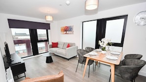 A modern living room with a sofa, dining table, and chairs. There is a TV, a plant, and a clock on the wall. - Spacious 2 Bed Apartment in Central Liverpool (Liverpool)