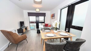 A modern dining area with a wooden table set for two, a sofa, and a TV in the background. - Spacious 2 Bed Apartment in Central Liverpool (Liverpool)