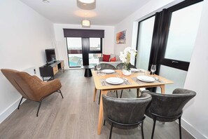A modern dining area with a wooden table set for two, a sofa, and a TV in the background. - Spacious 2 Bed Apartment in Central Liverpool (Liverpool)