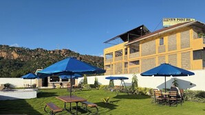 Un hôtel à la façade jaune, doté d’une terrasse avec des parasols bleus et offrant une vue sur un paysage vallonné.