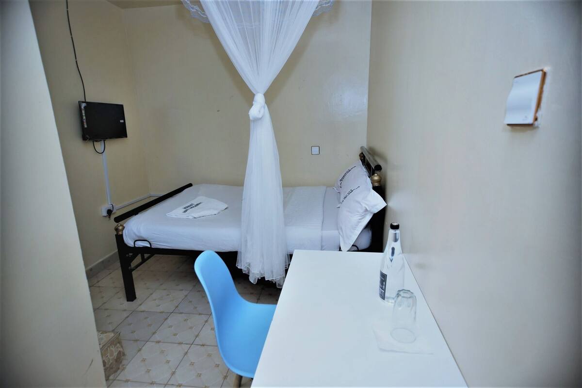 Una habitación pequeña con cama individual, mosquitera, mesita de noche, una silla y un escritorio con otra silla.