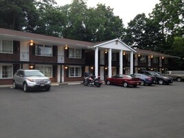 Un motel con entrada central, varios espacios de estacionamiento y fachada de ladrillo.