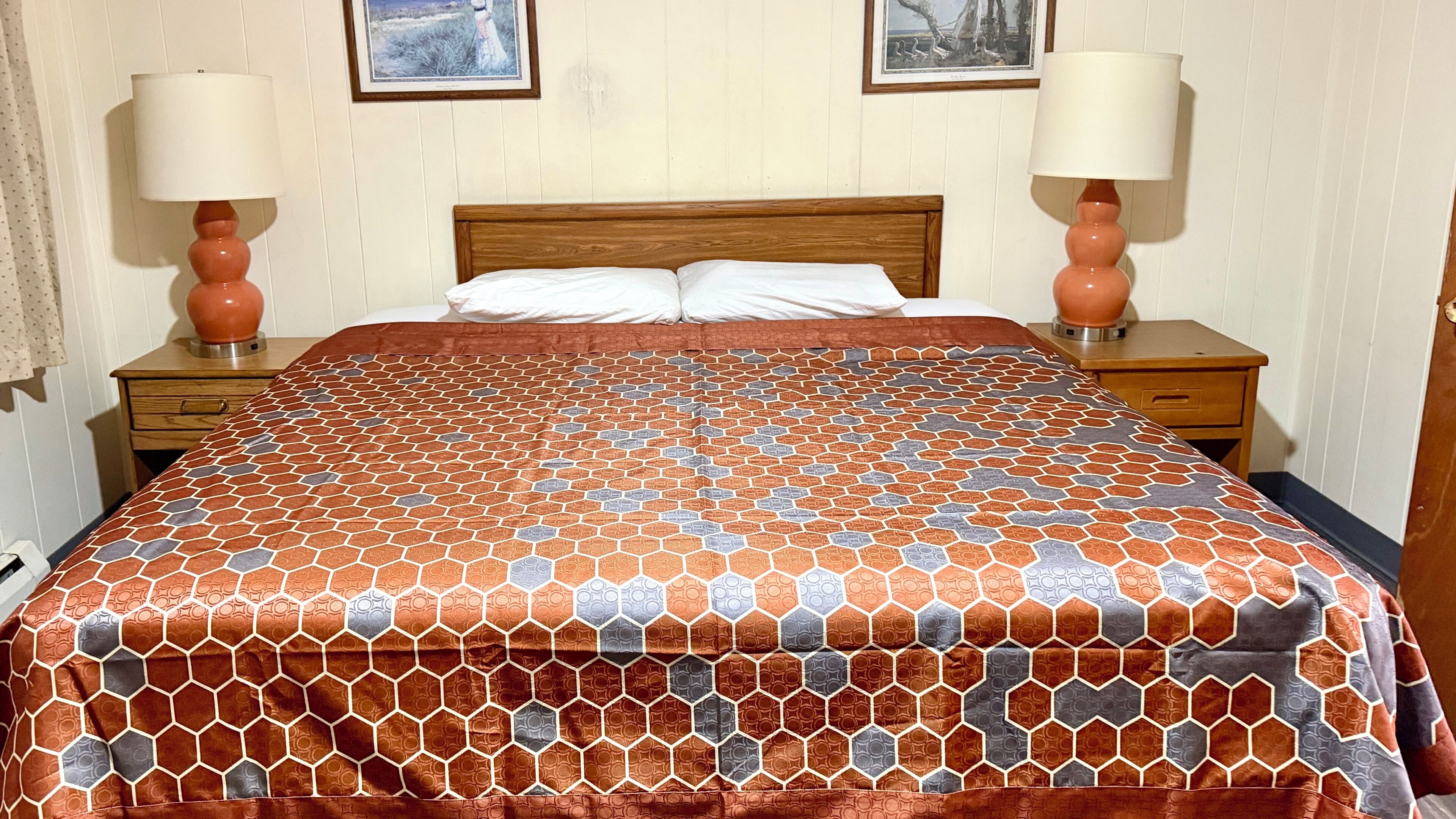 A hotel room with a wooden headboard, a patterned bedspread, two bedside lamps, and a nightstand with a drawer.