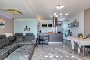 A modern living room with a grey sectional sofa, a dining table with chairs, a kitchen area with a stainless steel range hood, and a small island with a sink. - The Splendid Grande Pyramide with Sea View, Wi-Fi, and Air Conditioning (La Grande-Motte)
