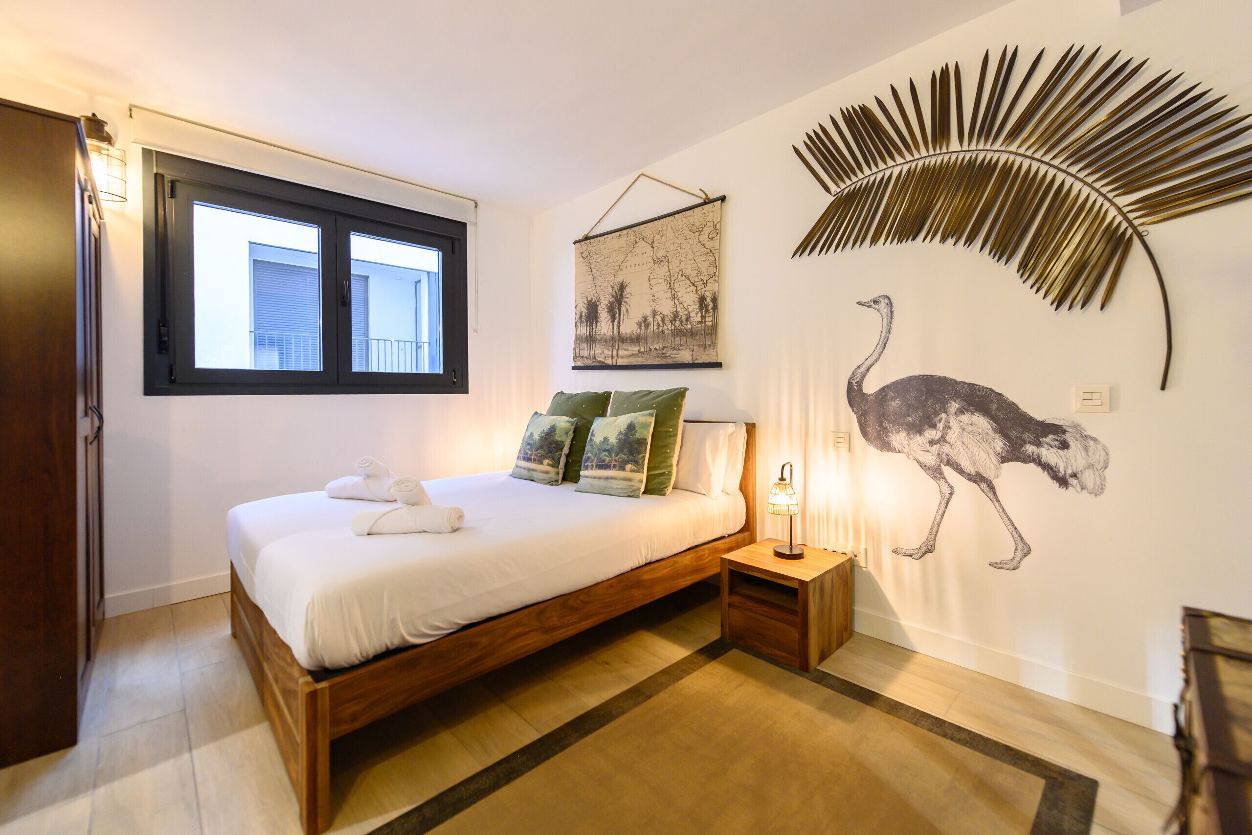 A bedroom with a bed, a nightstand, a wall-mounted lamp, a window, and a wall art of an ostrich.