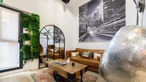 Ein modernes Wohnzimmer mit Couch und Kissen, einem Holztisch, einem Globus, einem Stadtdruck und einer grünen Wand mit Pflanzen.