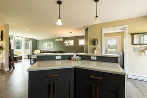 A kitchen with dark cabinetry, a large island, pendant lighting, and a view to a living area. - Morrison’s Beach Road Island Retreat (Georgetown)