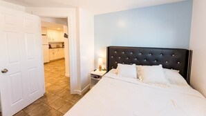 Hotellihuone, jossa on suuri sänky, yöpöytä lampun kanssa ja näkymä keittiöön avoimesta ovesta.