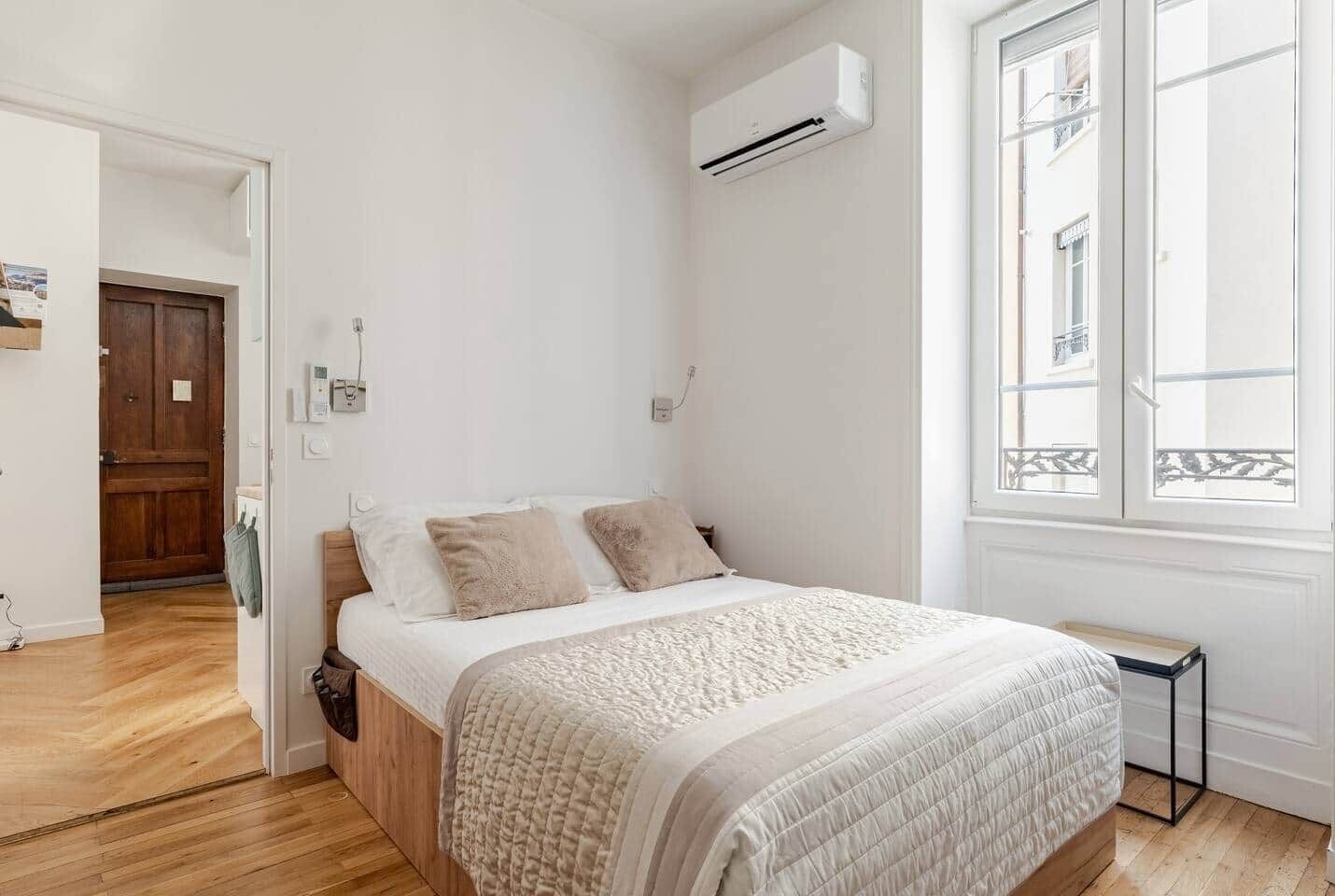 A bedroom with a bed, wooden floor, white walls, an air conditioning unit, a window, and a door.