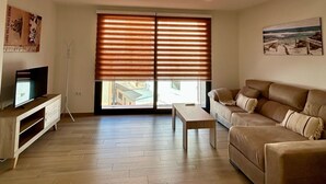A living room with a sofa, a TV on a stand, a wooden floor, and a large window with blinds. - Apartment in A Hotel 'Apartamento Type 2 A' with Private Terrace, Wi-Fi and Air Conditioning (Puente Genil)