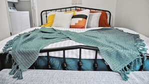 A neatly made bed with a teal blanket, a variety of pillows, and a wall-mounted laundry sign. - Boho abode Uptown Maumee by River-GREEN Side! (Maumee)