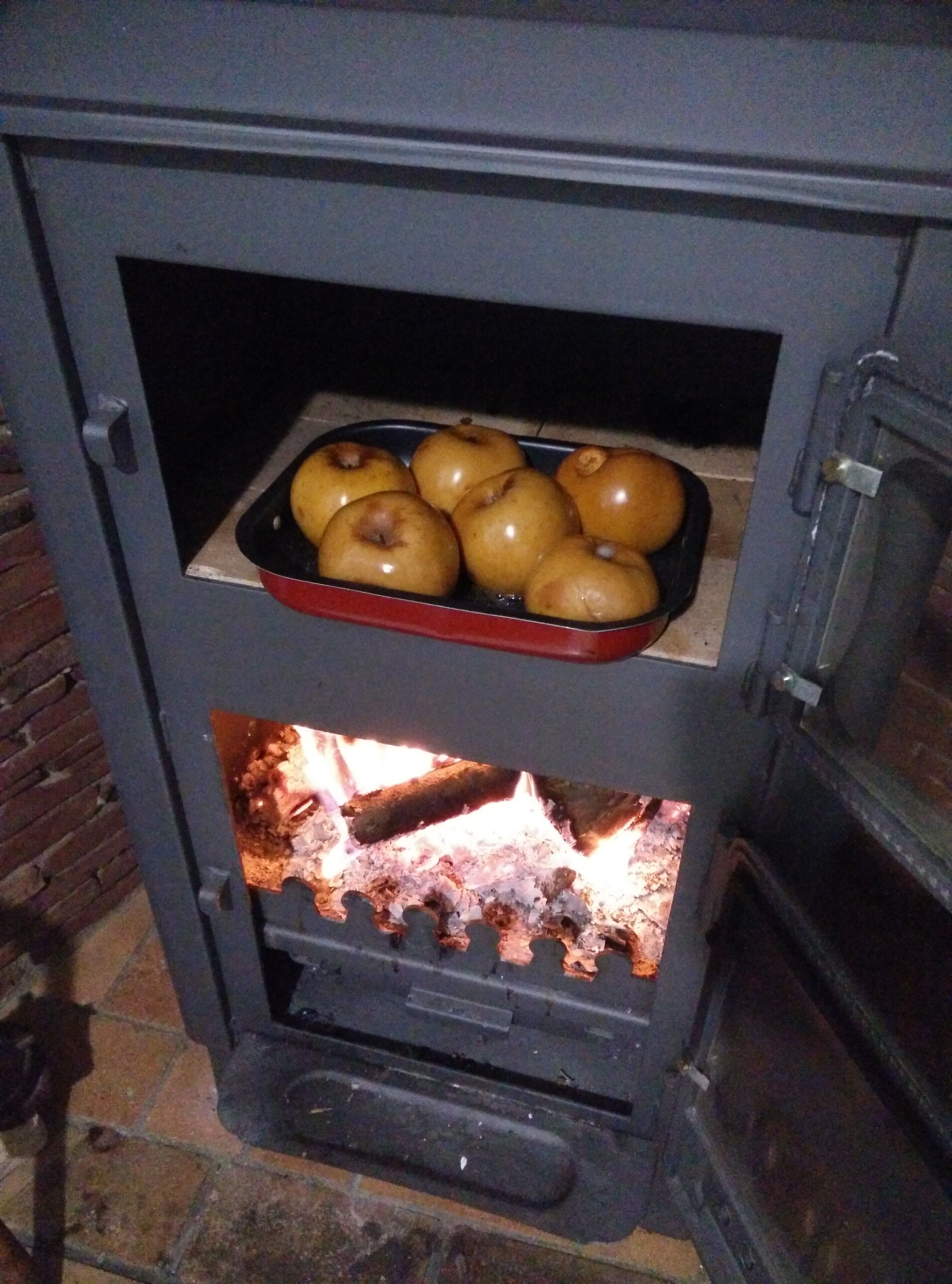 Un horno de leña con una bandeja de manzanas y fuego encendido en su interior.