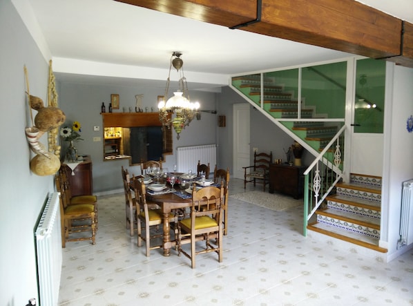 A dining area with a wooden table set for four, surrounded by chairs with yellow cushions. A chandelier hangs from the ceiling, and a staircase with green steps leads to an upper level.