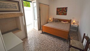 A bedroom with a bed, two bedside tables with lamps, a chair, a small table, a door leading to a balcony, and a framed picture on the wall. - Villetta Macao 1 in Torre Suda (Racale)