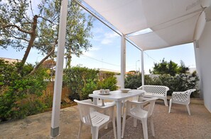 Een overdekt terras met een witte tafel en stoelen, omgeven door groen en een heldere lucht.