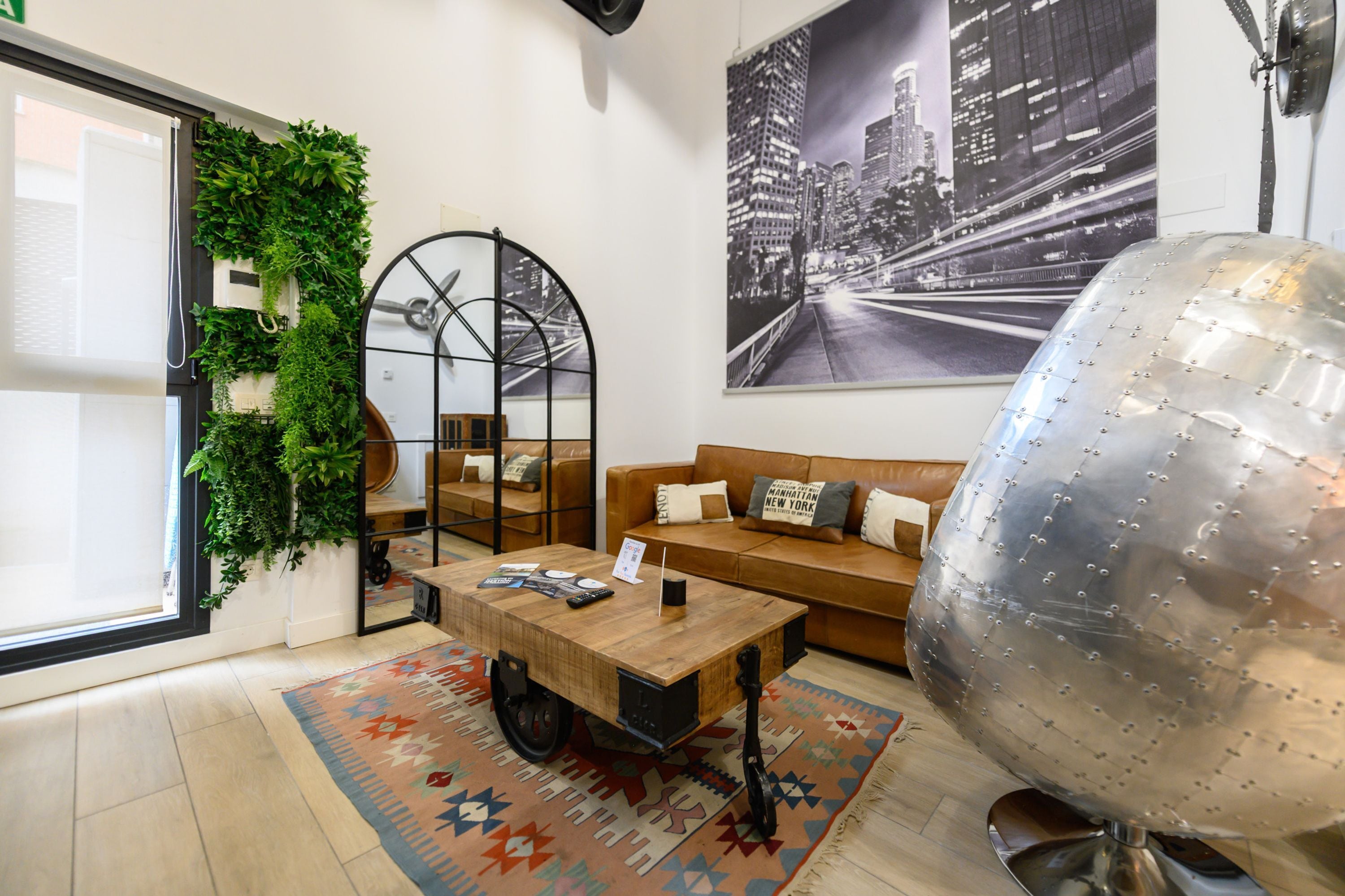 Una sala moderna con una mesa de centro de madera, un sofá con cojines, un globo metálico grande, un cuadro de un paisaje urbano y un jardín vertical verde.