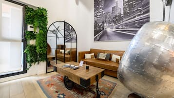 Una sala moderna con una mesa de centro de madera, un sofá con cojines, un globo metálico grande, un cuadro de un paisaje urbano y un jardín vertical verde.