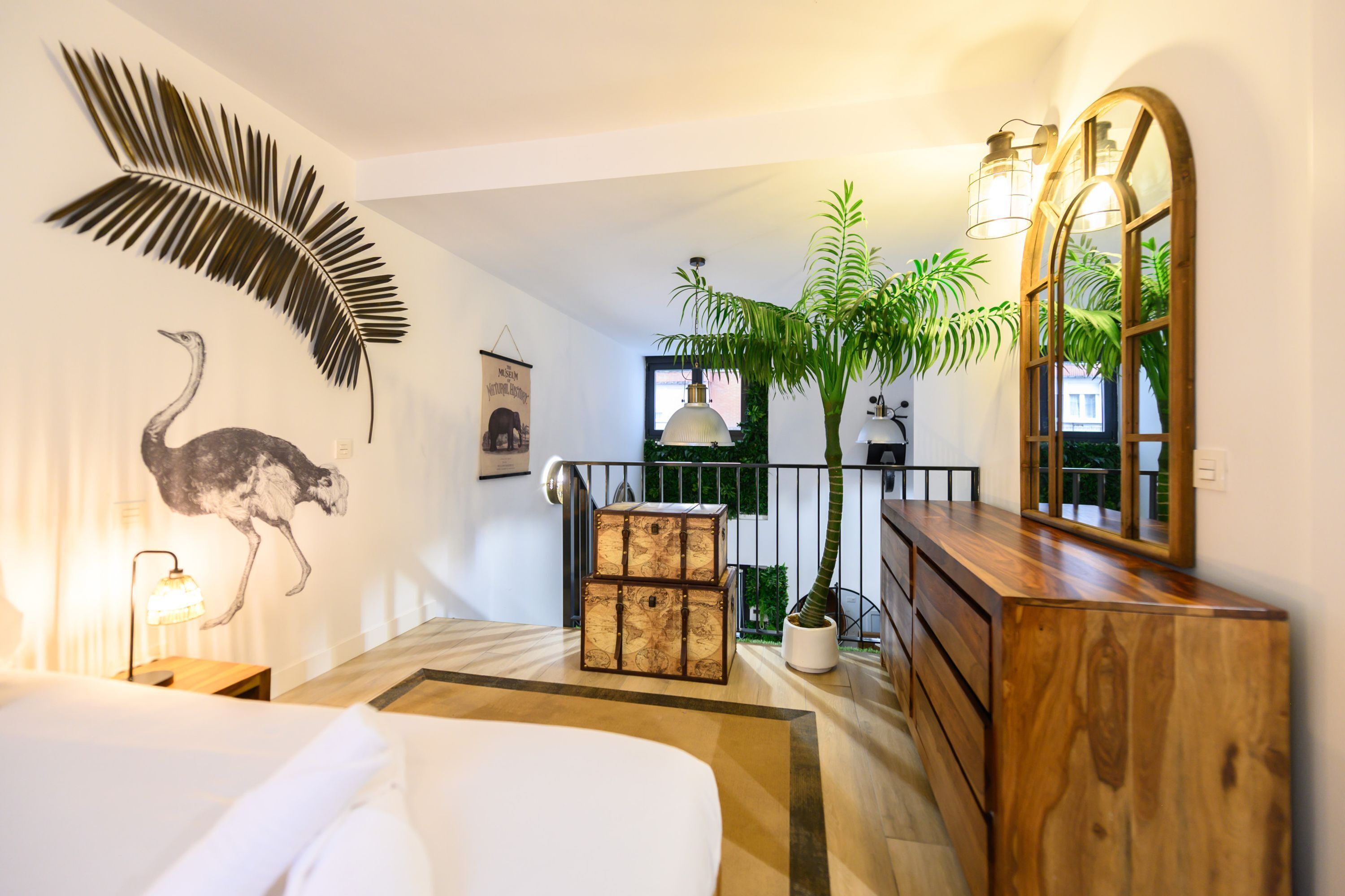 Habitación de hotel con cama, cómoda de madera, espejo, planta en maceta, cuadro de avestruz y adorno de pared.