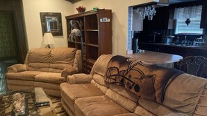 A living room with two beige sofas, a glass coffee table, and a small side table. There is a wooden bookshelf with decorative items and a sign that reads "KIN". A kitchen area is visible in the background with a dining table and chairs.