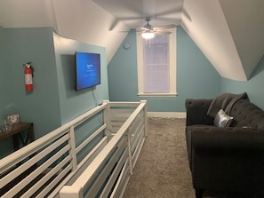Una habitación con paredes verde azulado, un ventilador de techo, un televisor montado, una ventana con persianas y un sofá oscuro, además de una cuna blanca.