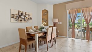 A dining area with a wooden table and chairs, a painting on the wall, and a view of the outdoors through large windows.