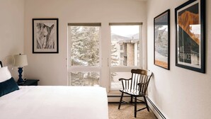 Ett hotellrum med en säng, en stol, en lampa och ett stort fönster med utsikt över träd och byggnader.