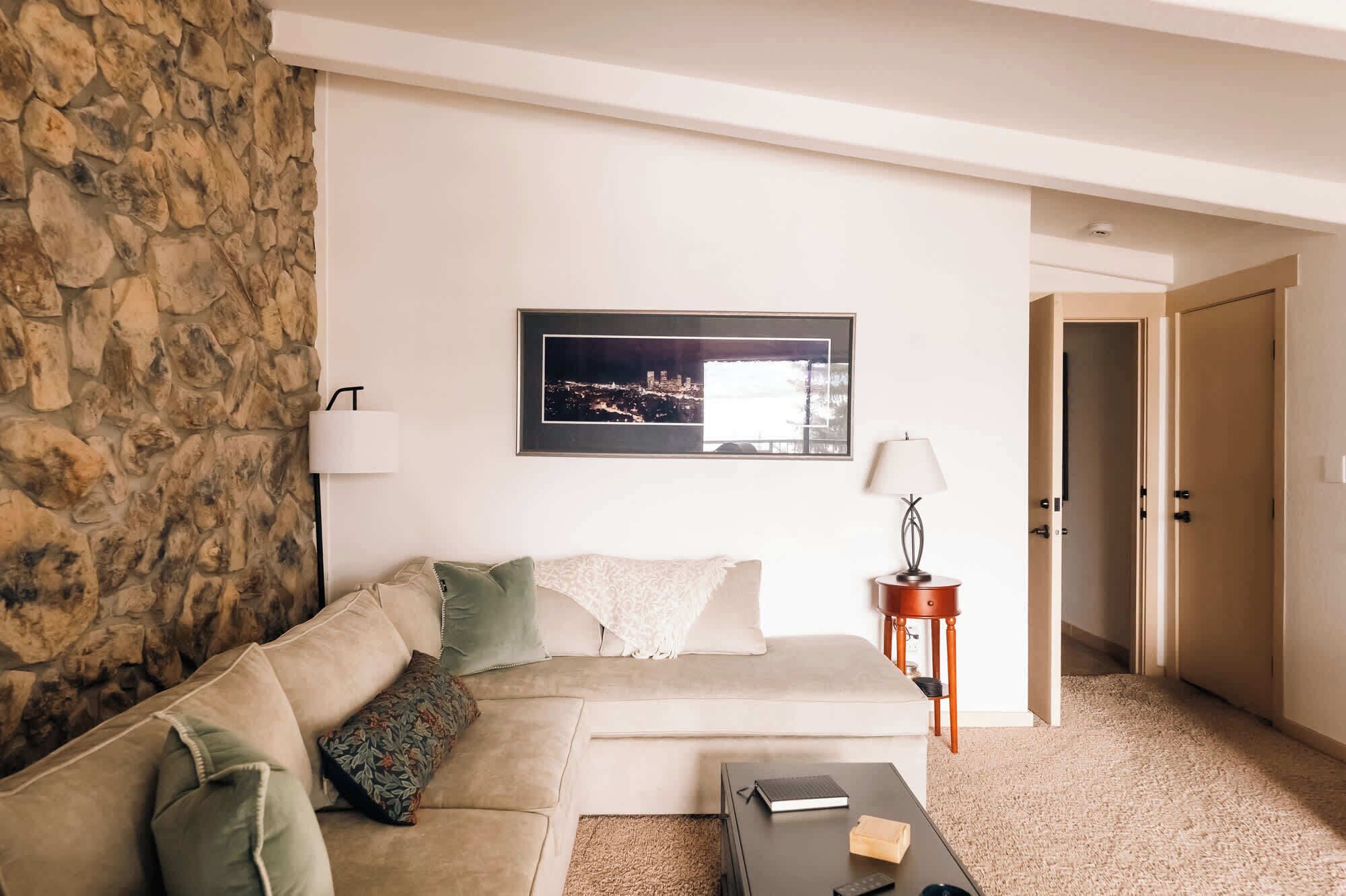 Un salon avec un mur en pierre, un canapé, une table basse, un tableau encadré représentant un paysage urbain, une lampe et une porte.